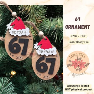 May include: Two wooden Christmas ornaments with the text "THE YEAR OF 67 2025". Each ornament has a red Santa hat design at the top. The ornaments are suspended on a string against a backdrop of green pine needles.