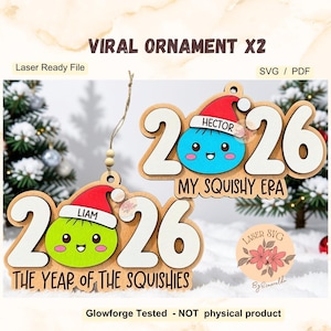Pode incluir: Dois ornamentos de madeira com o ano 2026 em branco e uma personagem colorida com um gorro de Pai Natal vermelho. Um ornamento apresenta o nome Liam e o texto THE YEAR OF THE SQUISHIES, o outro mostra Hector e MY SQUISHY ERA.