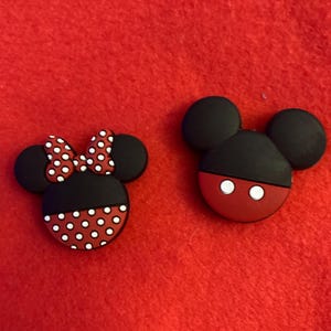 May include: Two cartoon character-themed shoe charms. One charm features a black head with a red base and white eyes. The other charm has a black head, a red base with white polka dots, and a red bow with white polka dots.