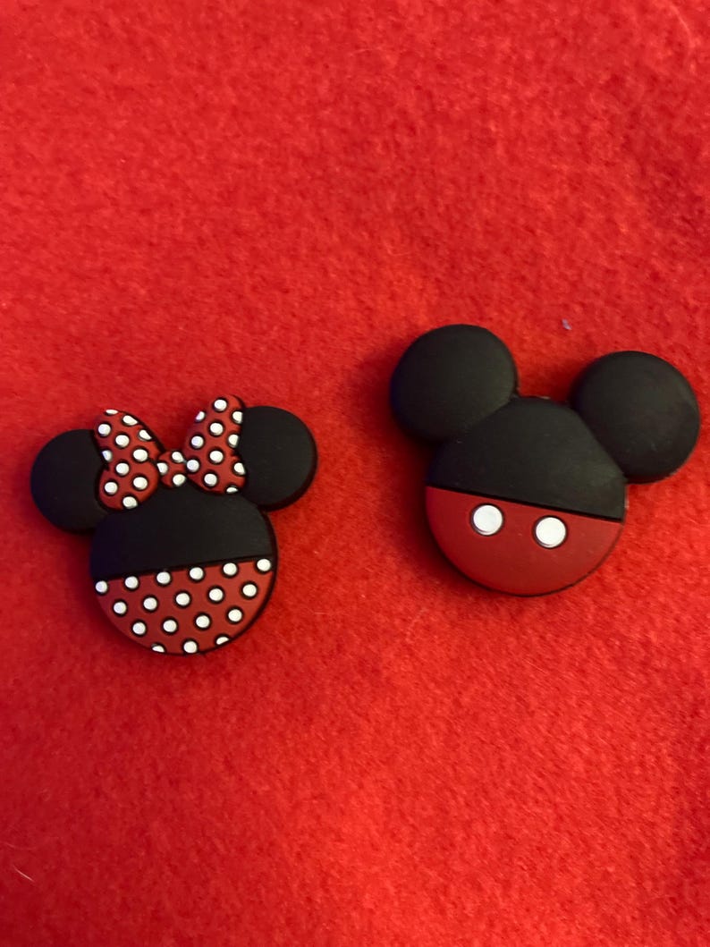 May include: Two cartoon character-themed shoe charms. One charm features a black head with a red base and white eyes. The other charm has a black head, a red base with white polka dots, and a red bow with white polka dots.