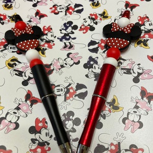 May include: Two pens with Minnie Mouse-themed toppers. One pen is black, the other is red with glitter. The toppers feature Minnie's iconic bow and ears. The background is a pattern of Minnie Mouse characters.