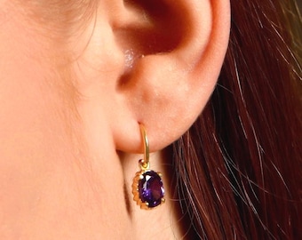 May include: A gold-colored earring with a purple oval-shaped gemstone. The earring is hanging from a person's ear. The gemstone is surrounded by a decorative gold setting.