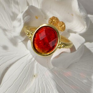 Puede incluir: Un anillo de oro con una piedra preciosa roja engastada en un engaste de bisel. El anillo está sobre una flor blanca.