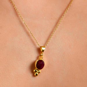 May include: A gold-colored necklace with a small, oval, dark red gemstone pendant. The pendant is accented with three small gold-colored beads below the gemstone. The necklace chain is delicate and appears to be made of gold.