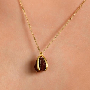 May include: A gold-colored necklace with a pendant featuring a dark brown gemstone. The pendant is encased in a gold-colored setting and hangs from a delicate gold chain. The necklace is displayed against a skin-toned background.