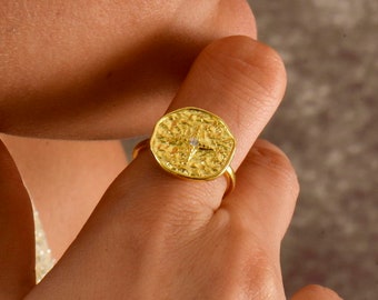 Puede incluir: Un anillo dorado con un diseño circular texturizado. El anillo se lleva en un dedo, mostrando sus intrincados detalles y su acabado brillante. El diseño del anillo presenta un elemento central, posiblemente una pequeña piedra o adorno.