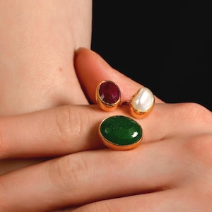 May include: Gold rings with gemstone settings. One ring features a large oval green stone, while the other has a red stone and a white pearl. The rings are displayed on a hand.