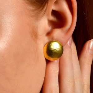 Könnte beinhalten: Nahaufnahme eines goldfarbenen Ohrrings mit gehämmertem Kugeldesign am Ohr. Ein goldener Ring mit einem kleinen, klaren Stein befindet sich an einem Finger. Der Hintergrund ist warmbraun.