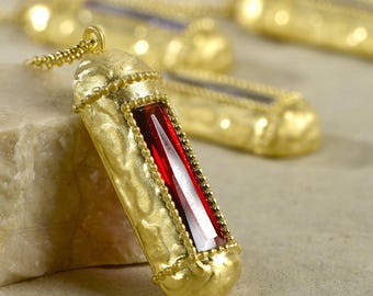 May include: A gold necklace with a pendant featuring a red gemstone set in a gold frame. The pendant has a textured surface and is surrounded by a beaded border.