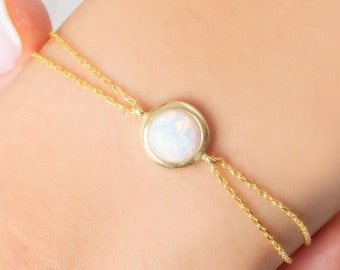 May include: A gold bracelet featuring a round, white opal stone set in a gold bezel. The bracelet has a delicate chain design, with two parallel strands extending from the opal. The jewelry is displayed on a person's wrist.