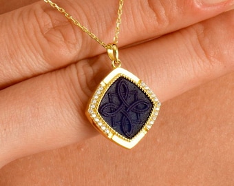 May include: A gold chain necklace with a square pendant featuring a black stone with a detailed design and a border of small clear stones.