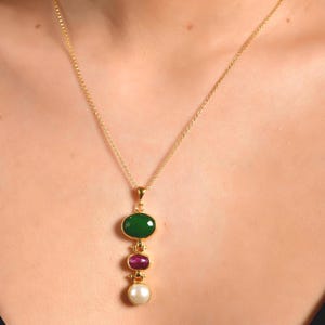 Könnte beinhalten: Eine goldfarbene Halskette mit einem Anhänger, der drei Edelsteine ​​aufweist: ein grünes Oval, ein rotes Oval und eine weiße Perle. Die Halskette wird um den Hals getragen.