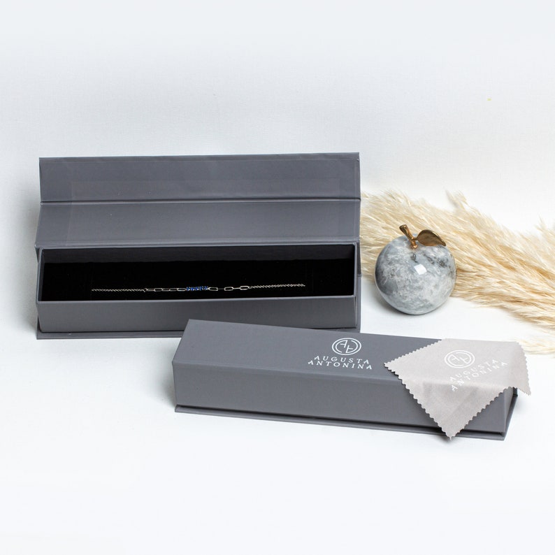 May include: A gray jewelry box with a silver bracelet inside. The box has the text "AUGUSTA ANTONINA" printed on the top. A gray cleaning cloth is next to the box.