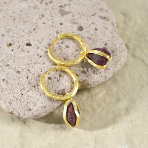 May include: A pair of gold-colored hoop earrings with a textured finish. Each earring features a deep red, oval-shaped gemstone. The earrings are displayed on a light gray stone surface.