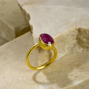 May include: A gold ring featuring a large, oval-cut ruby gemstone. The ruby is a deep, vibrant red color, set in a gold bezel. The ring's band is a simple, polished gold. The ring is displayed on a light-colored, textured surface.