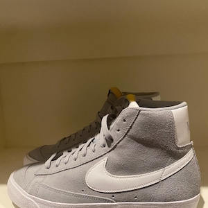 May include: Gray suede high-top sneakers with white soles and a white Nike swoosh. The sneakers have white laces and a padded collar. The shoes are displayed on a shelf.