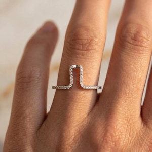 May include: A delicate silver ring with a unique architectural design. The ring features a straight bar that curves upwards into a rectangular shape, encrusted with small, sparkling stones. The band is also adorned with similar stones.