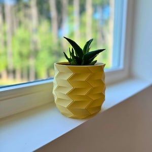May include: A yellow geometric planter with a small green succulent plant inside. The planter is sitting on a white windowsill.