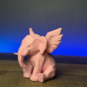 May include: A pink, geometric elephant figurine with stylized wings. The elephant is in a seated position, with its trunk curled and ears extended. The wings are detailed with individual feathers. The figurine is made of a solid material and has a matte finish.