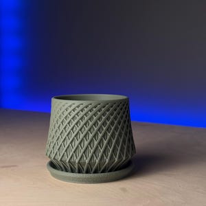 May include: A green geometric planter with a criss-cross pattern. The planter has a wide base and a narrow top.