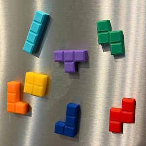 May include: A collection of colorful Tetris-shaped magnets in various colors, including blue, green, purple, yellow, orange, and red. The magnets are arranged on a metallic surface, showcasing the iconic game pieces.