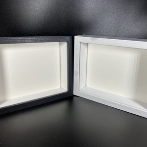 Puede incluir: Dos marcos de caja de sombra, uno negro y otro blanco, se muestran uno al lado del otro. Cada marco tiene una forma rectangular con un interior empotrado y un respaldo blanco. Los marcos son de madera y son adecuados para exhibir obras de arte u otros objetos.