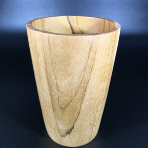 May include: A handcrafted wooden cup with a light brown finish. The cup has a tapered design, wider at the top and narrower at the base. The wood grain is visible, adding to its natural aesthetic. This cup is suitable for beverages.