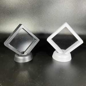 Könnte beinhalten: Zwei schwebende Vitrinen. Eine ist schwarz, die andere weiß. Jede Vitrine hat einen quadratischen Rahmen und eine transparente Mitte. Die Vitrinen sind auf einem kleinen, runden Sockel montiert, geeignet für Schmuck oder kleine Sammlerstücke.