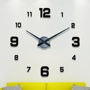 May include: A large, frameless wall clock featuring bold, black, three-dimensional numerals from one to twelve arranged in a circular pattern. The clock has a central circular hub with two long, slender, black metal hands indicating the time.