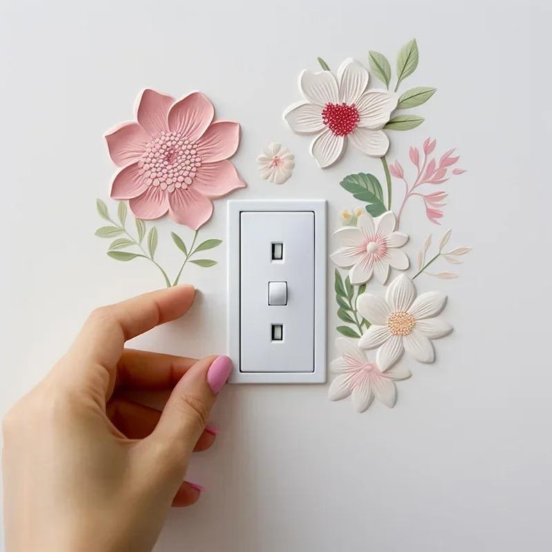 May include: A white light switch with two rectangular openings and a central toggle switch. The switch is surrounded by decorative, three-dimensional flowers in shades of pink, white, and green, creating a floral design.