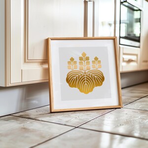 Puede incluir: Una impresión enmarcada con un diseño de cresta floral dorada sobre un fondo blanco. La obra de arte está en un marco de madera marrón claro, apoyada contra una pared. La imagen está sobre un suelo embaldosado.