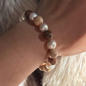 May include: A beaded bracelet featuring an alternating pattern of smooth white spherical pearls and textured brown wooden beads. The beads are strung together in a circular arrangement, showcasing a contrast between the lustrous white surfaces and the dark, grained wood finish of the spherical components.