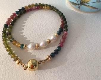 Könnte beinhalten: Ein mehrfarbiges Edelstein-Armband mit einem goldenen Verschluss und einer Reihe weißer Perlen. Das Armband besteht aus grünen, rosa und gelben Edelsteinen.