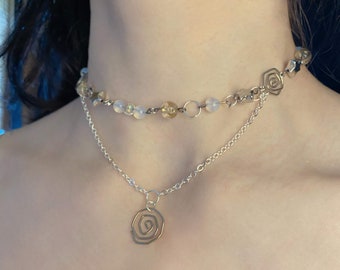 Puede incluir: Un collar de cadena de plata con un delicado colgante de rosa y una cadena de cuentas superpuesta con cuentas blancas y doradas.