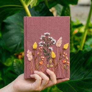May include: A rectangular notebook with a dusty rose fabric cover featuring intricate embroidery of various wildflowers and grasses in shades of white, yellow, and orange, with green stems and leaves detailed across the front surface.