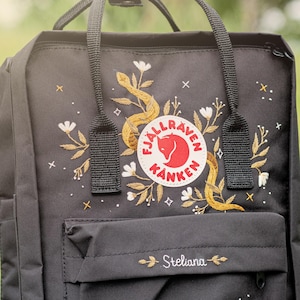 Puede incluir: Mochila negra con diseño bordado de flores y serpiente. Presenta un logotipo redondo y el nombre "Steliana" bordado en el bolsillo delantero. Incluye asa superior y correas ajustables.