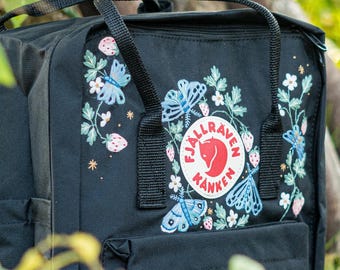 Può includere: Zaino nero con ricami di farfalle, fragole e fiori. Lo zaino presenta un manico superiore, spallacci regolabili e una tasca frontale. Il logo del marchio è una volpe rossa all'interno di un cerchio bianco.