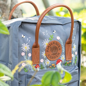 May include: A grey backpack with brown leather handles and embroidered floral details. The design includes white daisies, pink buds, red mushrooms, and bees. The backpack features a circular leather patch with the brand name.