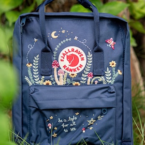 Puede incluir: Mochila azul marino con diseños bordados de flores y setas. La mochila presenta el texto "be a light for all to see" y un logotipo circular. Tiene un asa superior y un bolsillo delantero.