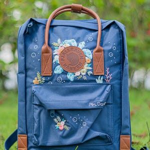 May include: A navy blue backpack with brown leather details. The front pocket has the text "Mrs. Davi". The backpack features a floral design with the brand logo. The bag has a top handle and adjustable straps.