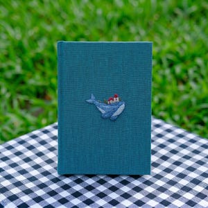 May include: A teal-colored book with a textured fabric cover. Centered on the front cover is a small, detailed embroidery featuring a light blue whale with a tiny red and white lighthouse perched on its back, rendered in fine thread work.