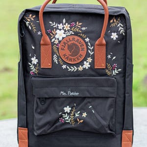 Könnte beinhalten: Schwarzer Rucksack mit braunen Lederdetails und Griffen. Mit gesticktem Blumendesign und rundem Logo. Auf der Vordertasche ist der Name "Mrs. Fletcher" eingestickt.