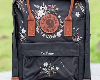 Può includere: Zaino nero con dettagli e manici in pelle marrone. Presenta un design floreale ricamato e un logo circolare. La tasca frontale ha il nome "Mrs. Fletcher" ricamato.