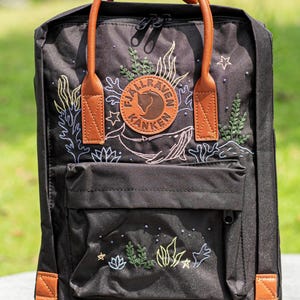 Op de afbeelding: Zwarte rugzak met bruine leren details en een handvat. De voorkant heeft een ronde patch met de merknaam en een walvisillustratie. Geborduurde ontwerpen van planten, sterren en waterleven versieren de tas.