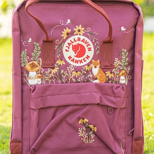 May include: A burgundy backpack with embroidered floral and woodland designs. The backpack features a fox, mushrooms, sunflowers, and the brand logo. It has a front pocket and burgundy handles.