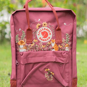 Op de afbeelding: Bordeaux rugzak met geborduurde bloemen- en bosontwerpen, waaronder zonnebloemen, paddenstoelen en een vos. De rugzak heeft een voorvak, een handvat aan de bovenkant en het "Fjallraven Kanken" logo.