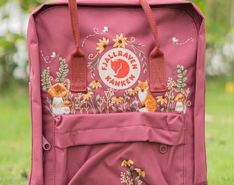 Può includere: Zaino bordeaux con ricami floreali e boschivi, tra cui girasoli, funghi e una volpe. Lo zaino presenta una tasca frontale, una maniglia superiore e il logo "Fjallraven Kanken".