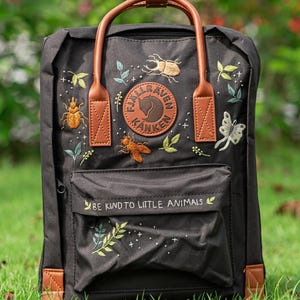 May include: Black backpack with brown leather accents and embroidered insects and leaves. Features a front pocket with the text "BE KIND TO LITTLE ANIMALS" and a circular logo. Includes a top handle and adjustable straps.