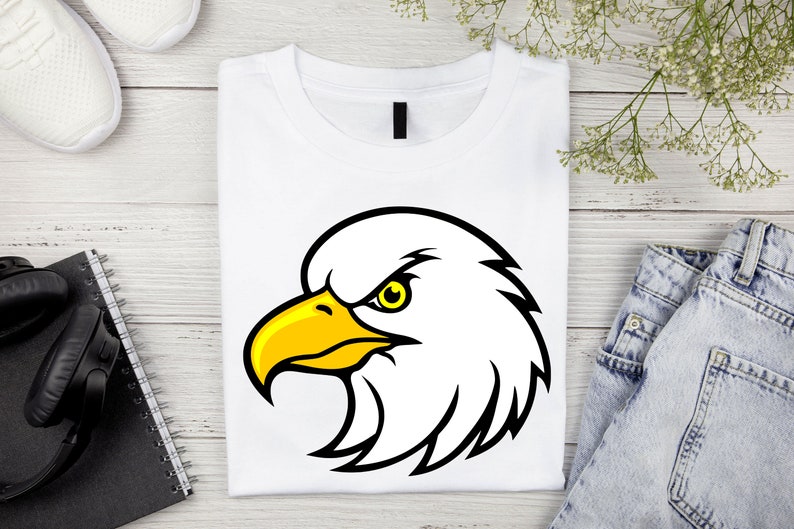 May include: White t-shirt with a black and yellow cartoon eagle head graphic. The eagle has a fierce expression with a yellow beak and a black eye.