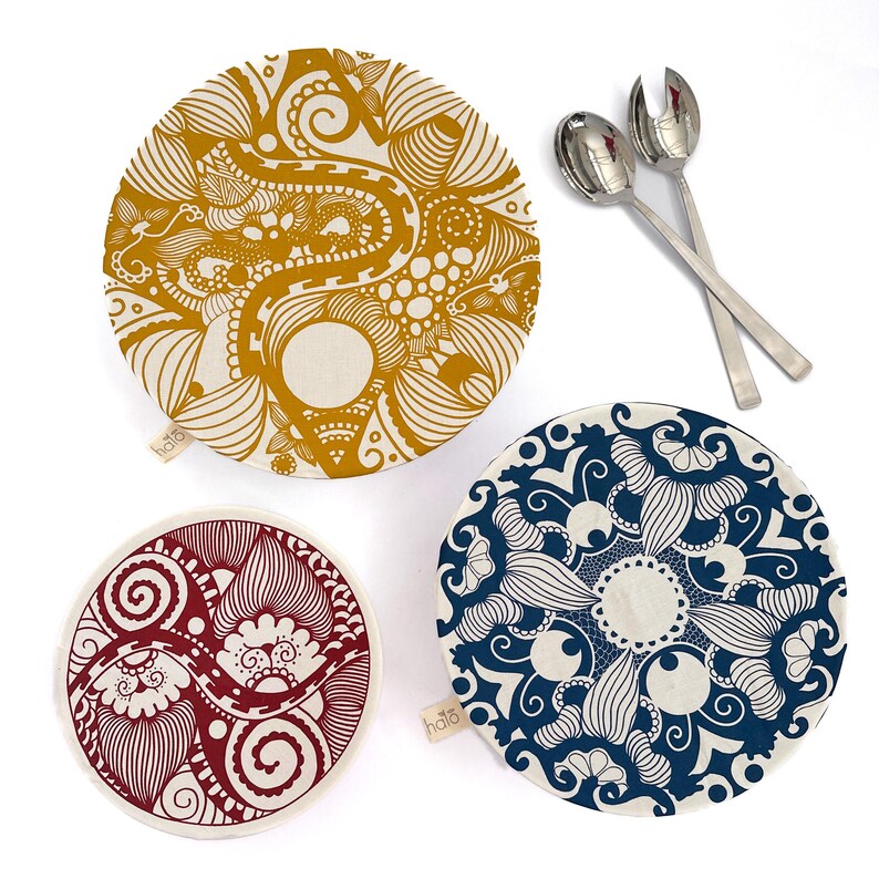 May include: Three reusable food covers with different patterned designs. The covers are made of fabric and have a white background with colorful patterns. The largest cover has a yellow and orange pattern, the medium cover has a blue pattern, and the smallest cover has a red pattern. The covers are labeled "hailo" in the bottom left corner.